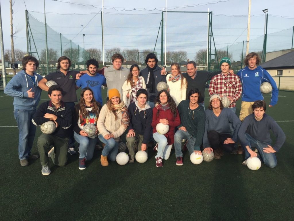 Group of 18 people on a soccer field with balls, smiling and posing for a group photo on a sunny day.