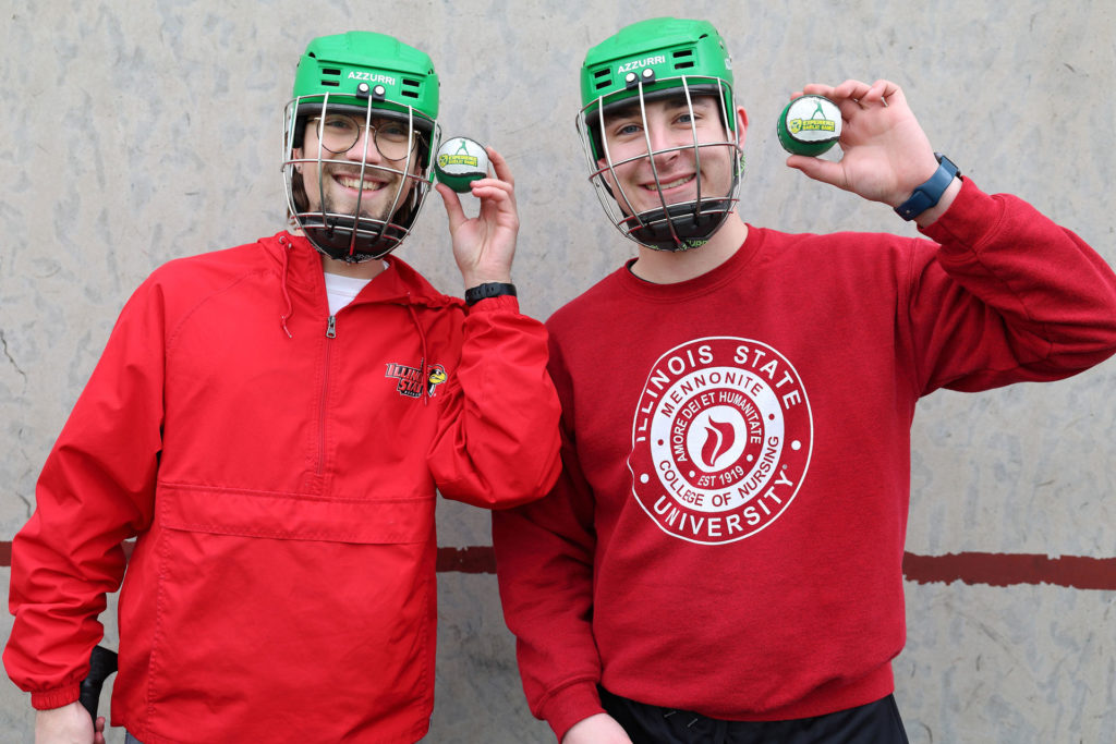 Two people in red Illinois State University gear hold lacrosse balls, smiling and wearing green helmets.