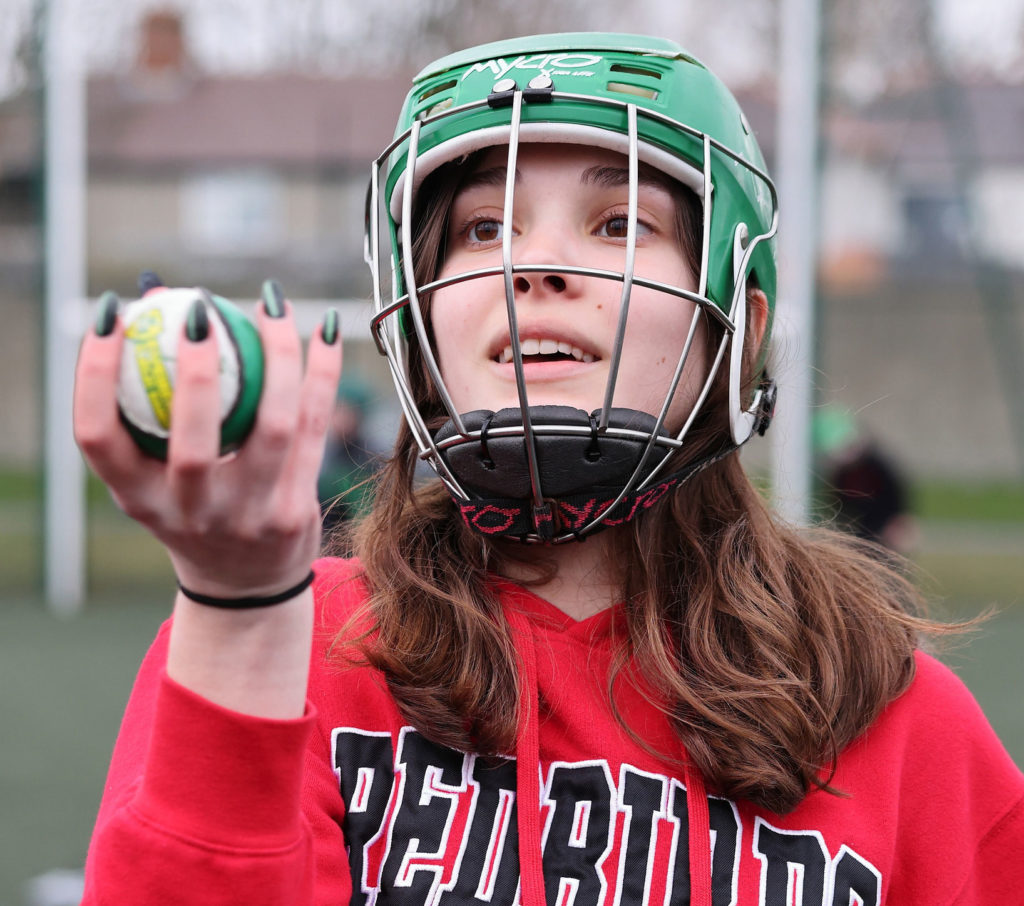 Young athlete in a green helmet holding a ball during a sports practice, wearing a red hoodie outdoors.