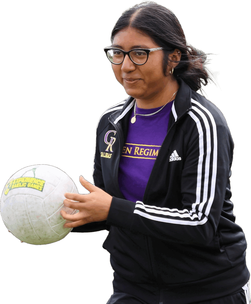 Person with glasses holding a Gaelic football outdoors, wearing a jacket and purple shirt.