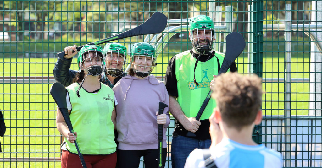 Group playing hurling outdoors, wearing green helmets and holding hurleys, posing for a photo at a pitch fence.
