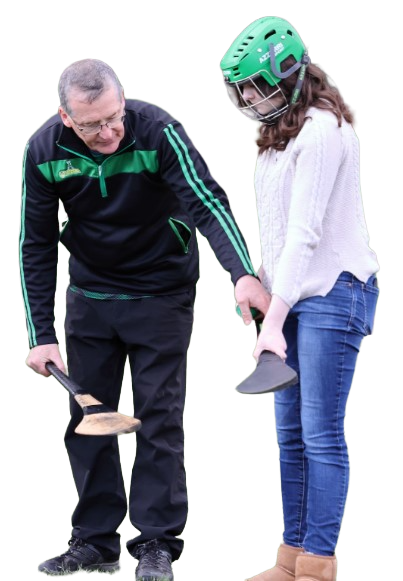 Coach instructs young player in hurling technique, both holding hurleys; player wears a green helmet for safety.