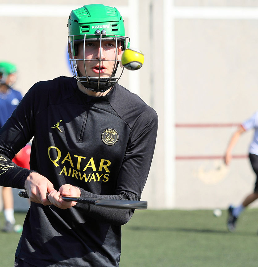 Hurling player focused on striking the ball during practice, wearing protective helmet and sports gear.