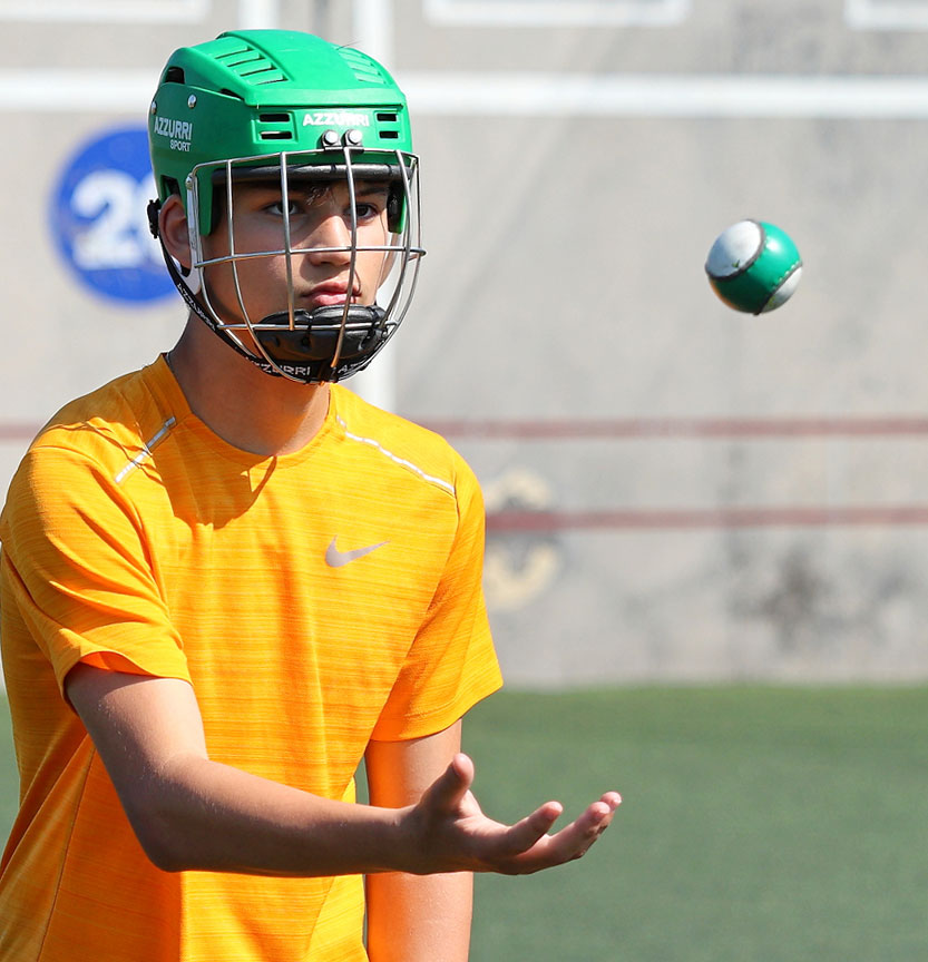 Teen in green helmet practicing pelotari skills with a ball, wearing an orange shirt on a sunny day.