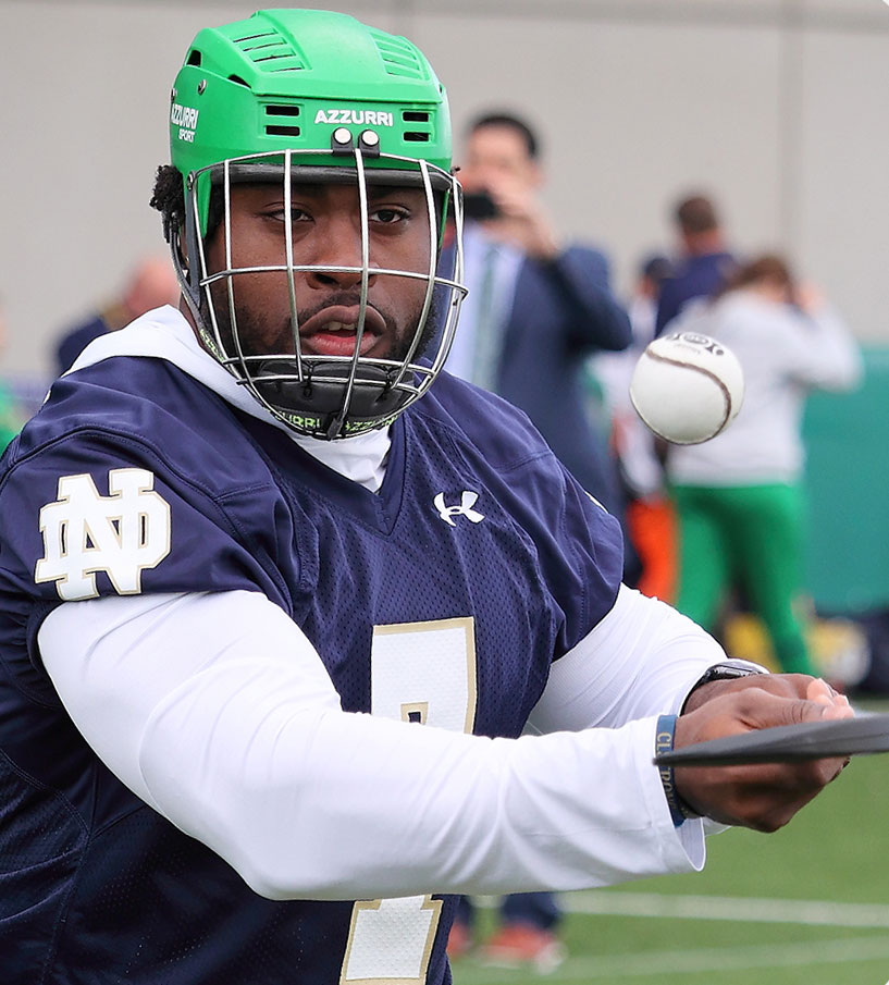 Notre Dame athlete practicing with a lacrosse stick in green helmet and blue jersey during training session.