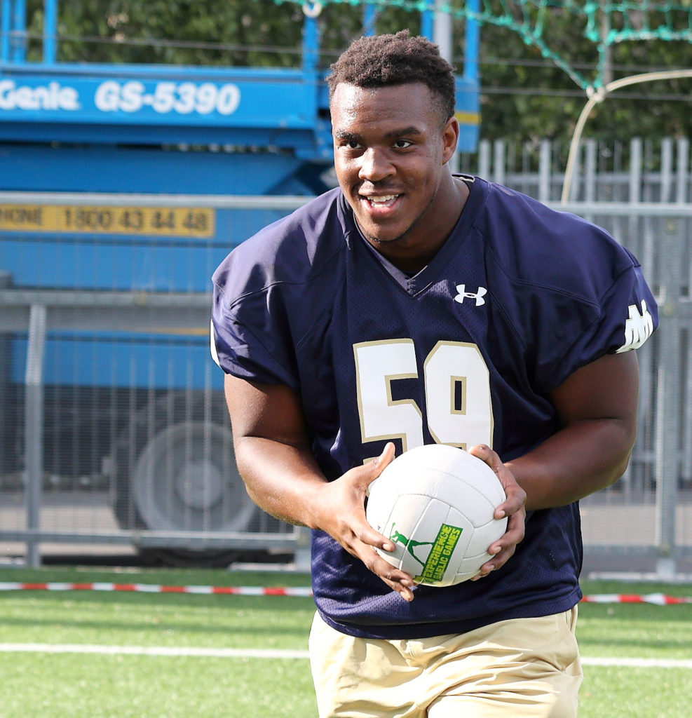 Football player holding a volleyball on a field, wearing a dark jersey with number 59, smiling outdoors.