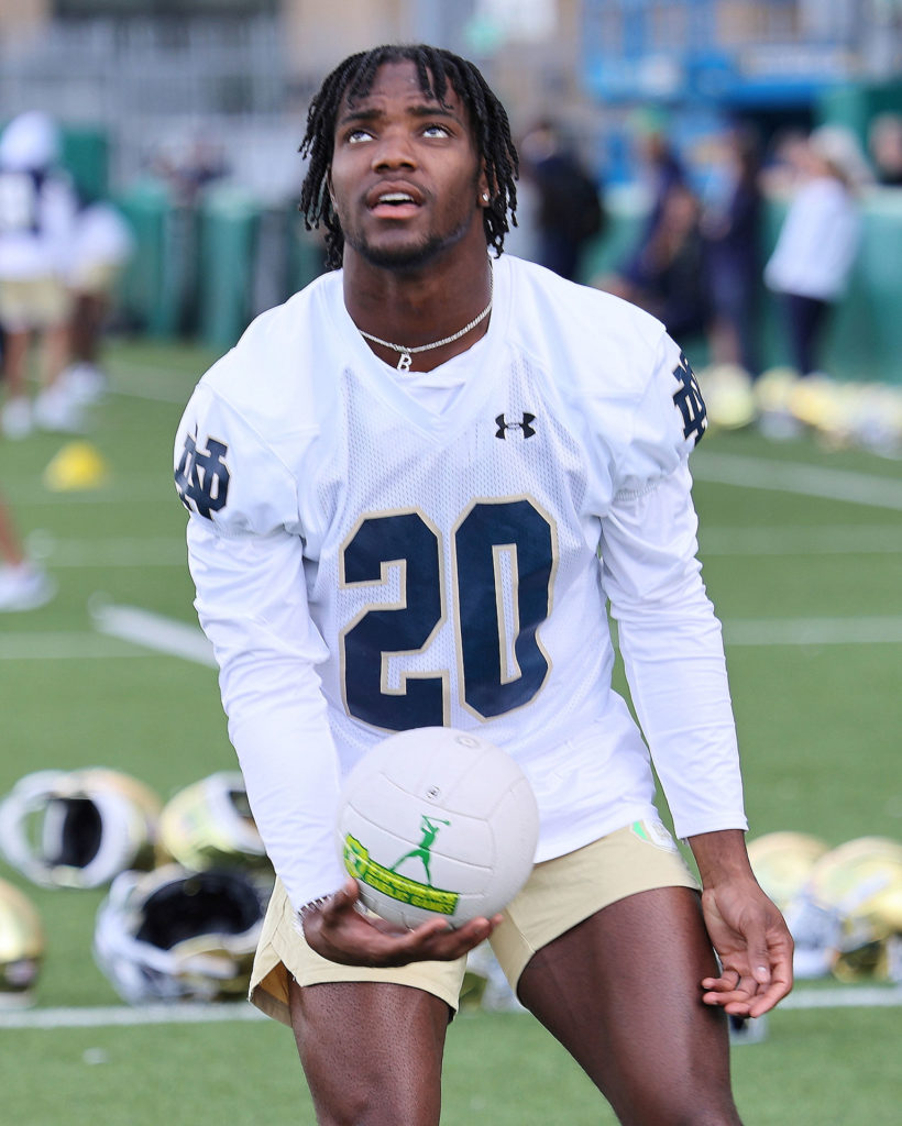 Football player in white jersey with the number 20 holding a volleyball during practice on a field.