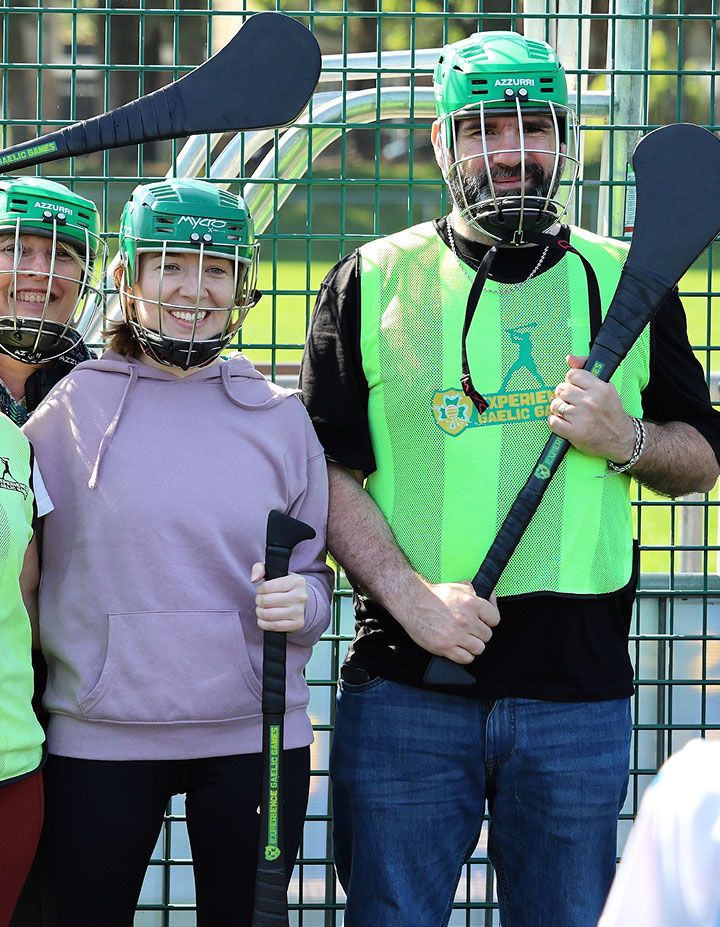 Two people in green helmets holding hurleys, ready for Gaelic games on a sports field.