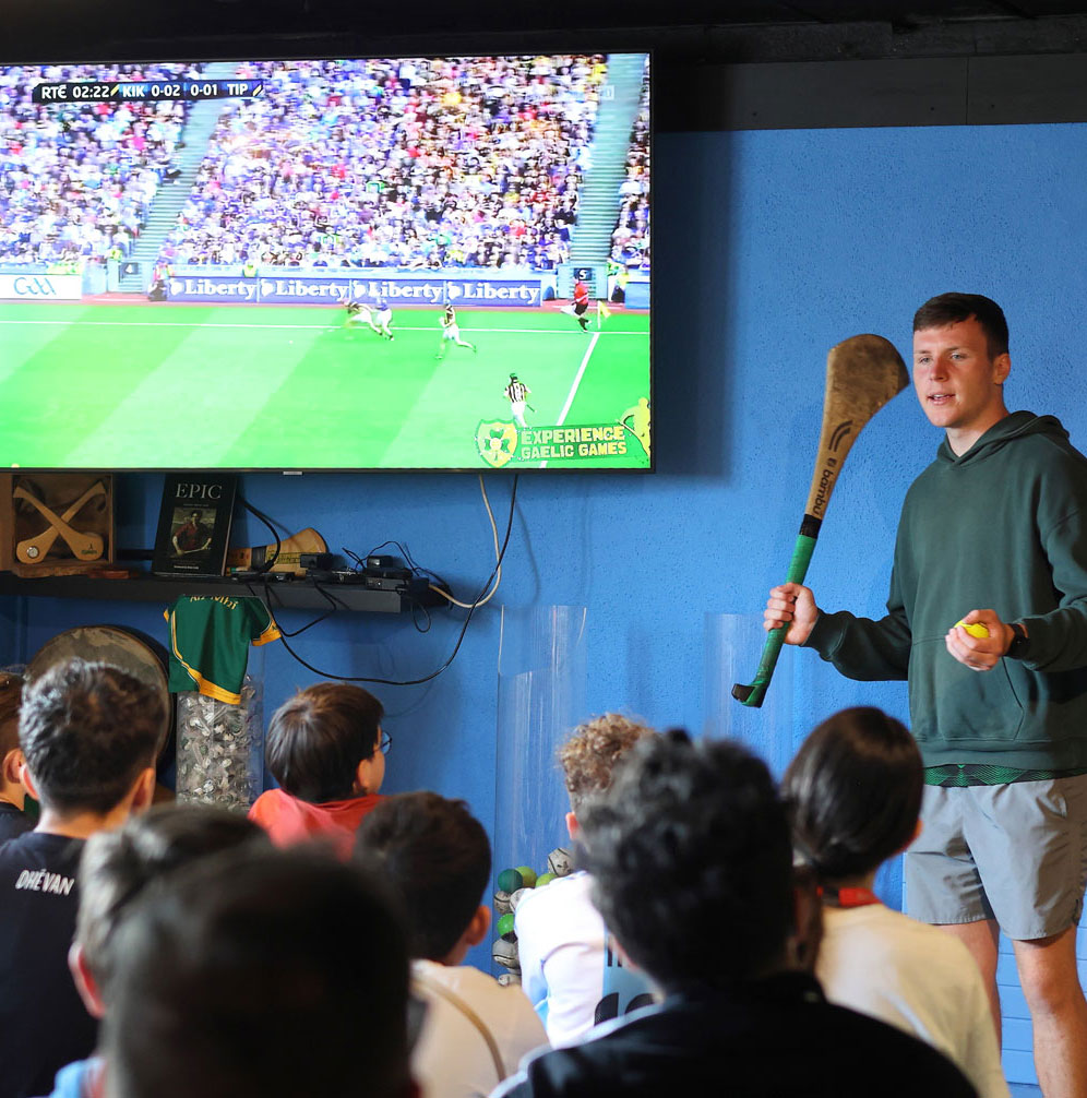 Instructor teaching Gaelic games to a group, with match shown on a TV screen in the background.
