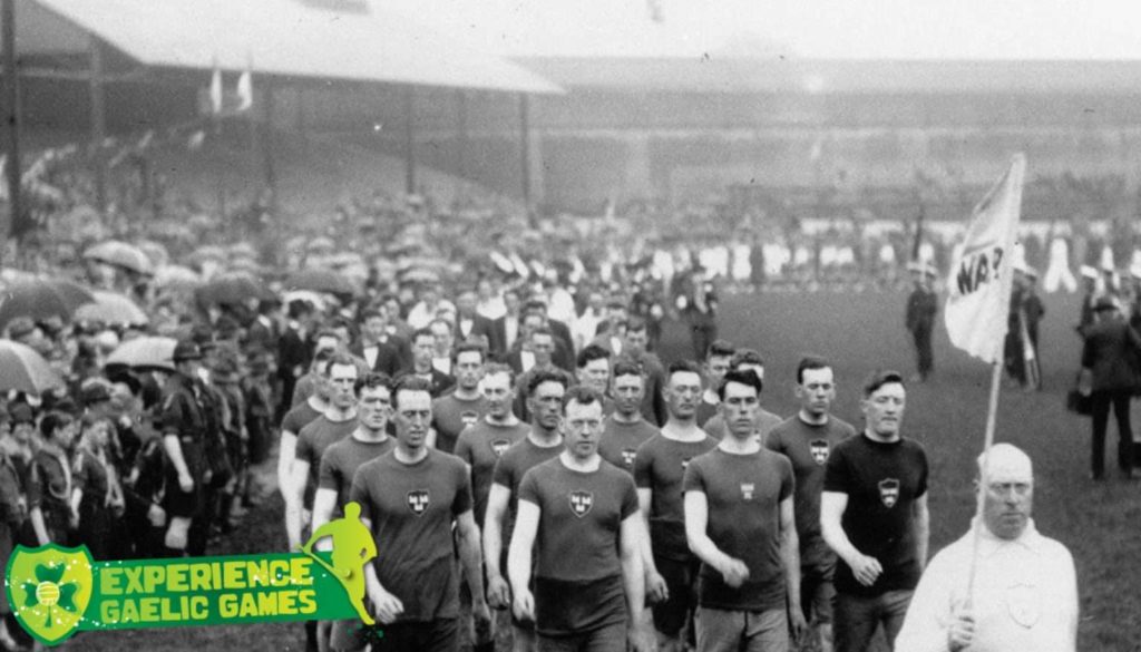 Vintage Gaelic Games athletes marching on a rainy day, with a crowd in the background and a flag bearer leading.