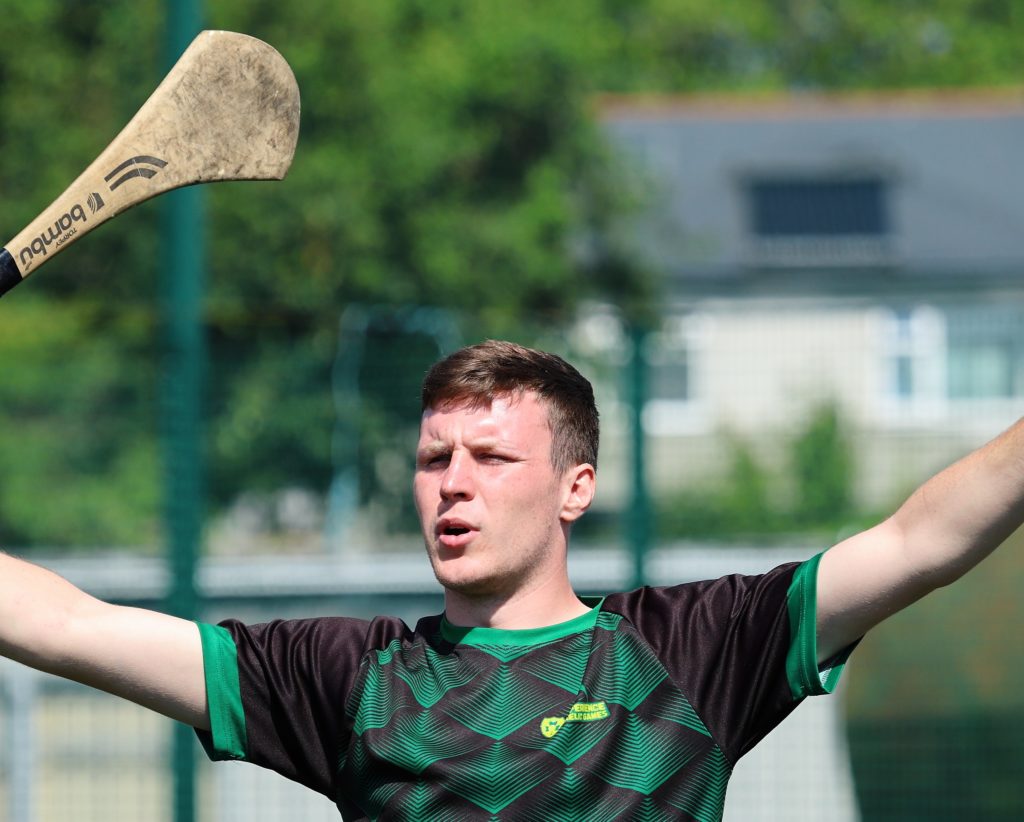 Man playing hurling on a sunny day, holding a hurley stick and raising arms on the field.
