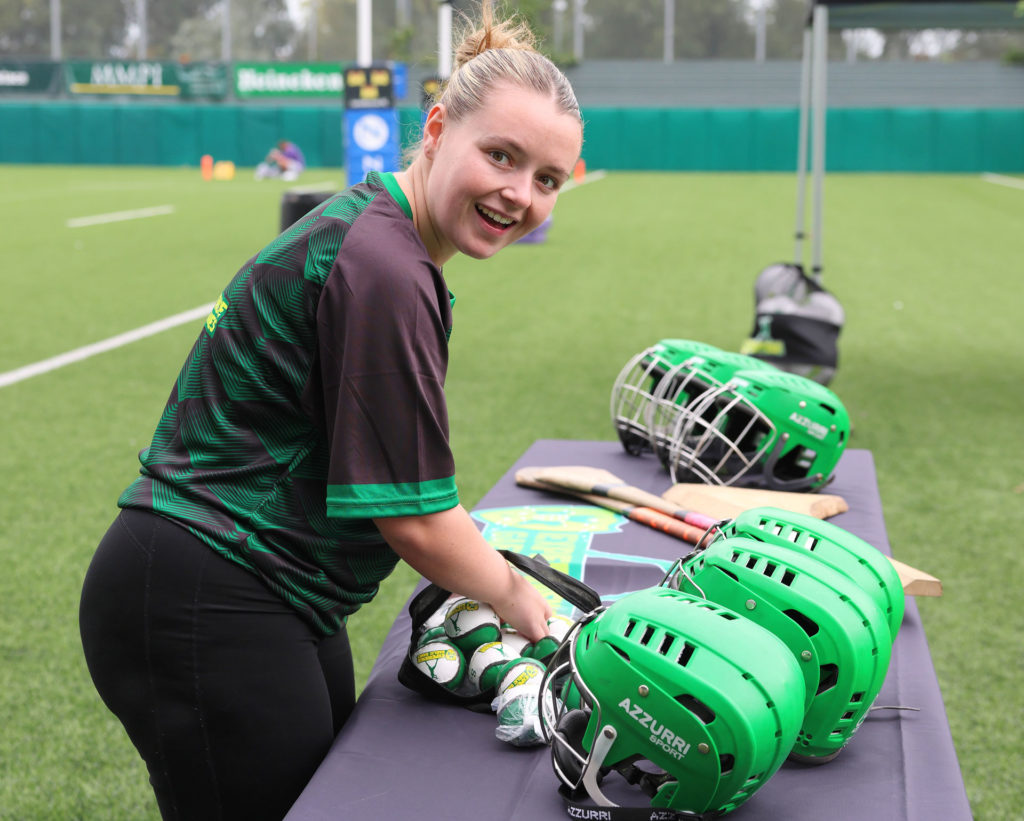 Smiling woman preparing hurling gear on a sports field, with green helmets and hurls on the table nearby.