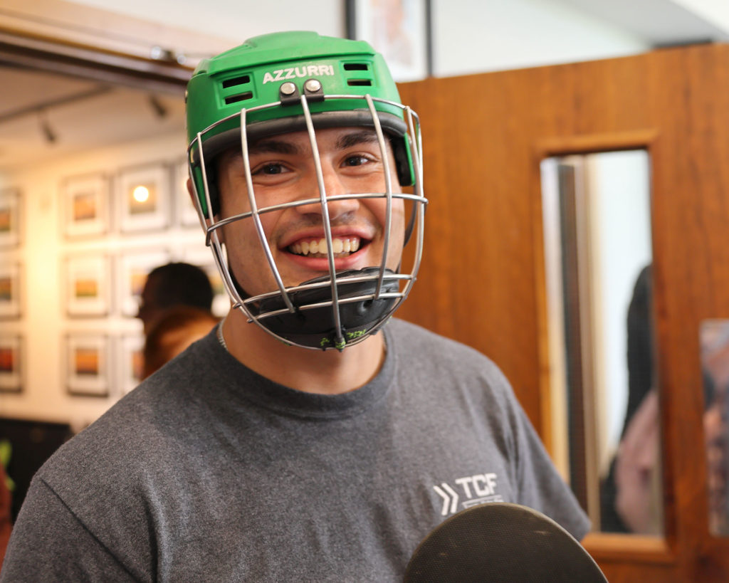 Smiling person wearing a green helmet and grey shirt indoors, holding a paddle.