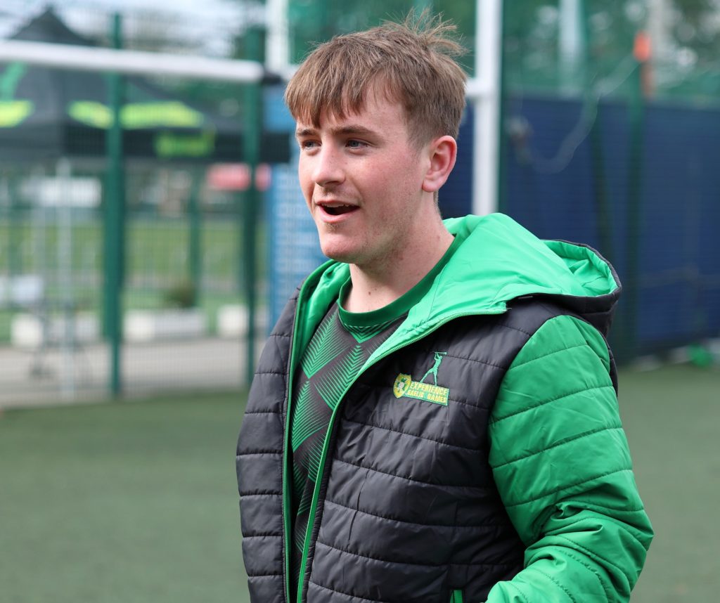 Young man in green jacket smiling outdoors on a soccer field.