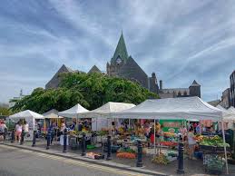 Outdoor market with white tents and fresh produce near historic church under a blue sky.
