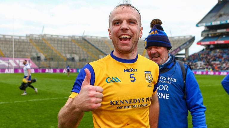 A happy athlete in a yellow jersey gives a thumbs-up on the field, with a smiling man in a blue hat in the background.