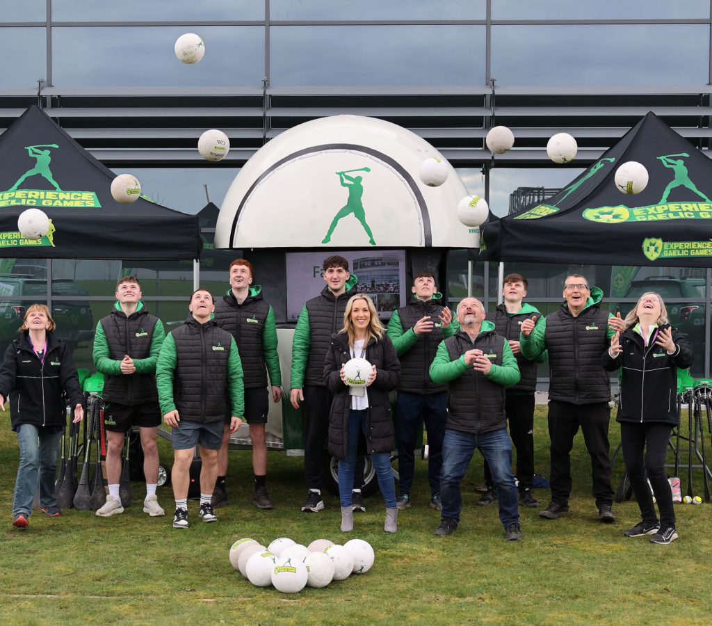 Group celebrating Gaelic games event, smiling team tossing balls outdoors in branded jackets under tents.