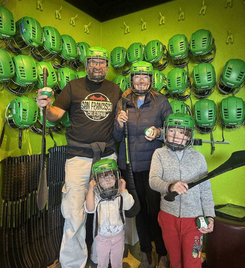 Family wearing green helmets and holding gear, ready for hurling game in a vibrant, themed room.