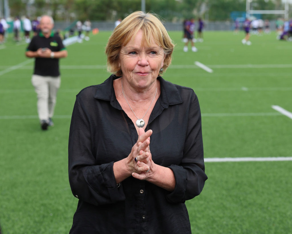 Woman standing on sports field smiling, with blurred background of people and greenery.