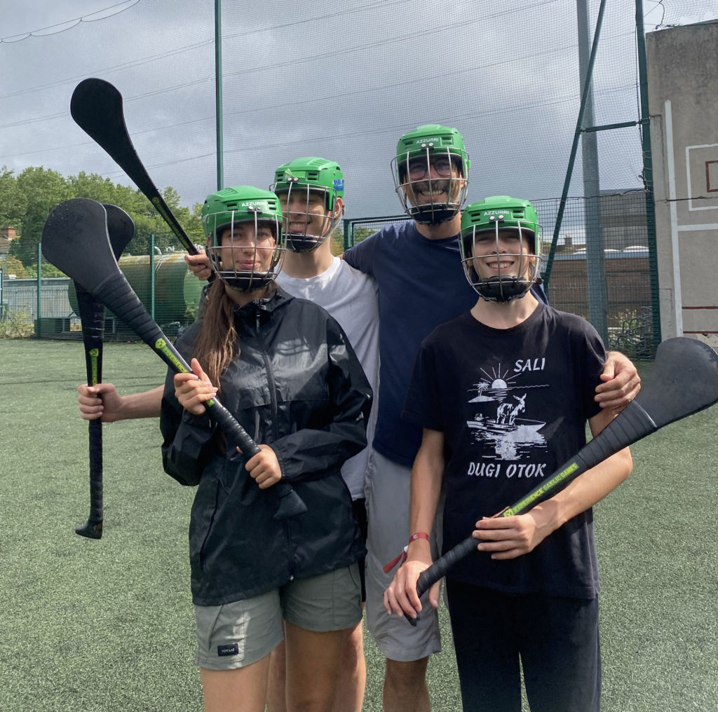 Four people in helmets and sports gear smiling on a field with hurleys, ready to play hurling.