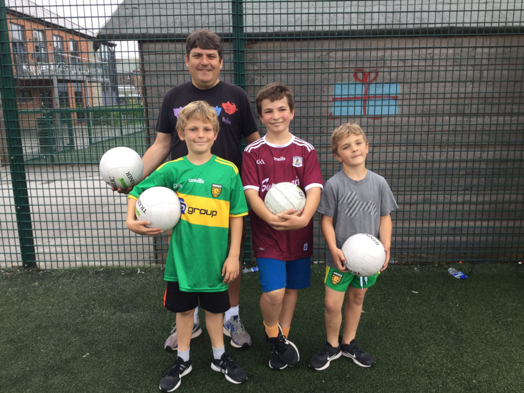 Man and three children holding footballs on a sports field near a fence, wearing colorful sports jerseys.