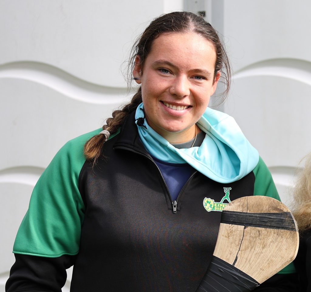 Woman holding a hurling stick, wearing a green and black sports jacket, smiles outdoors.
