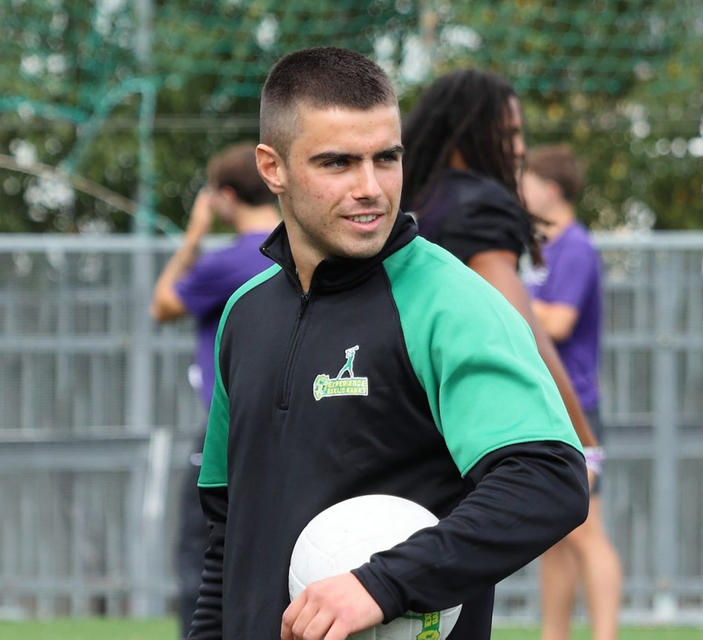 Man in a green and black sports jacket holding a soccer ball, outdoor sports field in the background.