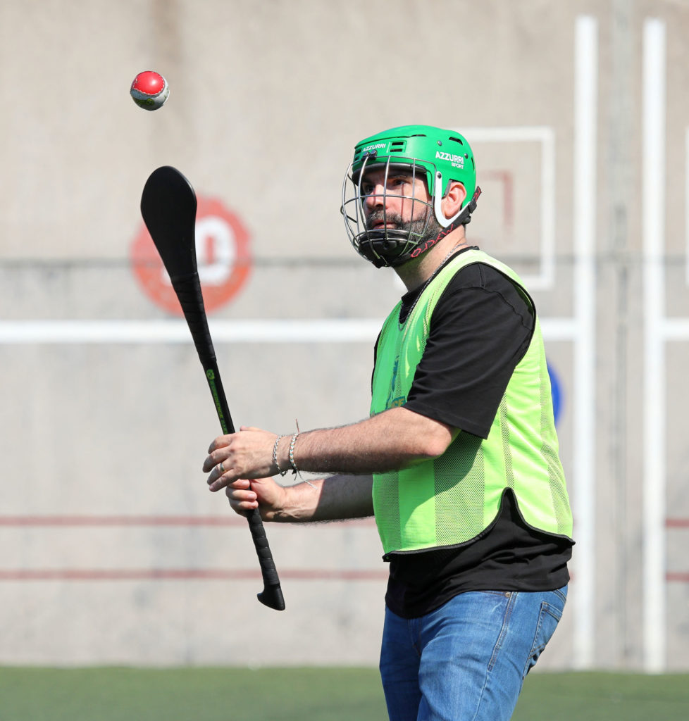 Man wearing green helmet plays hurling, focusing on a ball mid-air, on a sports field.