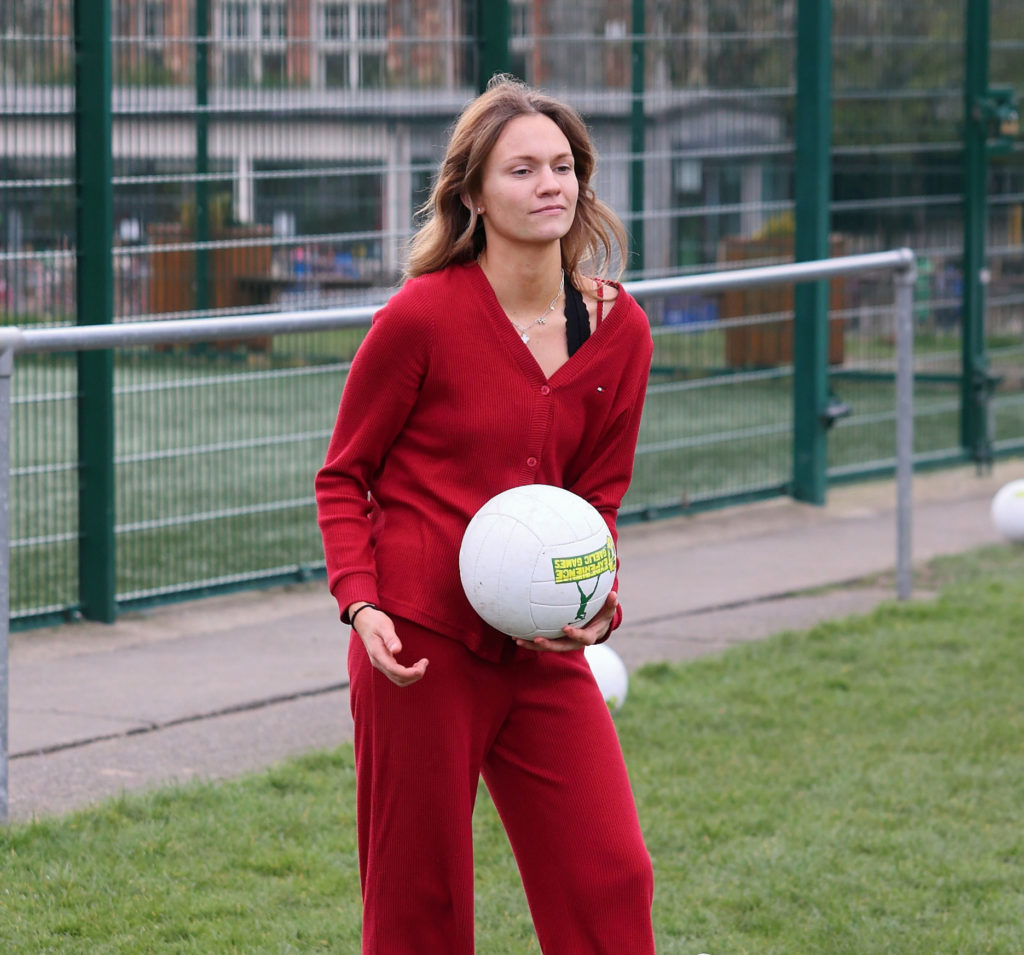Woman in red outfit holding a volleyball on a sports field, near a fenced area.