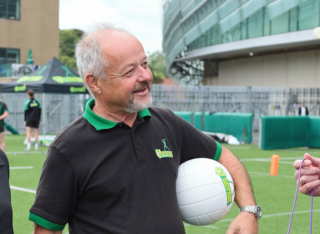 Man smiling on sports field, holding a volleyball, wearing uniform with a logo. Athletic activity in outdoor setting.