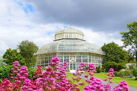 Victorian glass greenhouse surrounded by vibrant pink flowers and lush greenery under a cloudy sky.