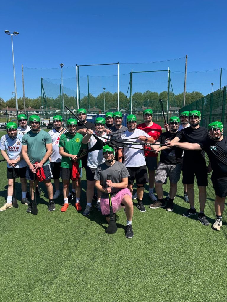 Group of men in sports gear on a field, wearing green helmets and holding hurling sticks under a clear blue sky.