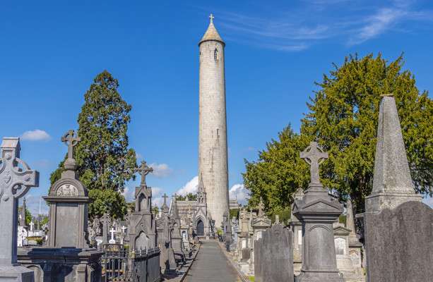 Cemetery landscape with tall round tower under blue sky, surrounded by trees and gravestones.