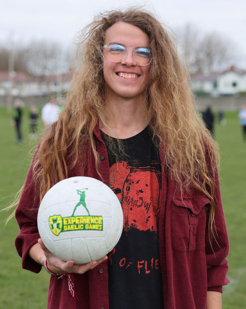 Smiling person holding Gaelic football, outdoors on a field.
