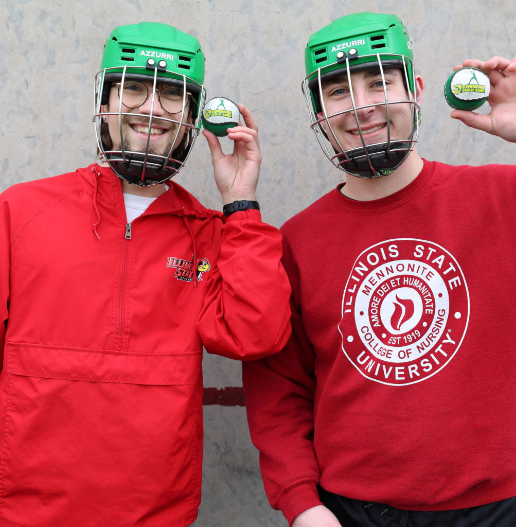 Two smiling men in green helmets holding hockey pucks, wearing Illinois State University jerseys.