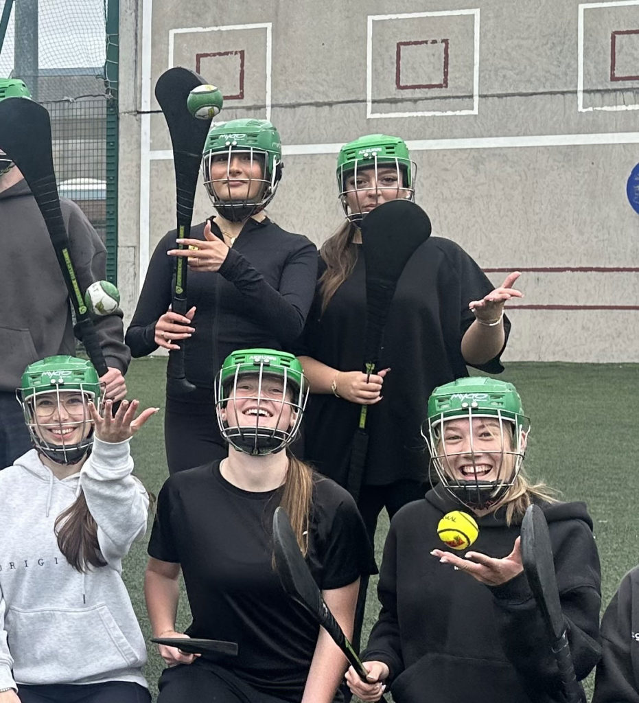 Women playing camogie, smiling and juggling sliotars while wearing helmets on a sports field.