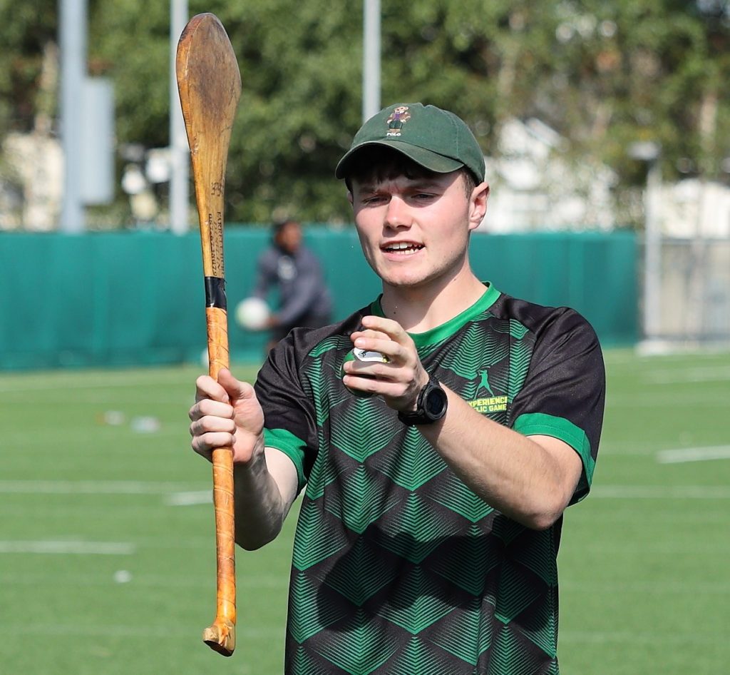 Young man in sports attire holding a hurling stick, preparing to play on a sunny field.