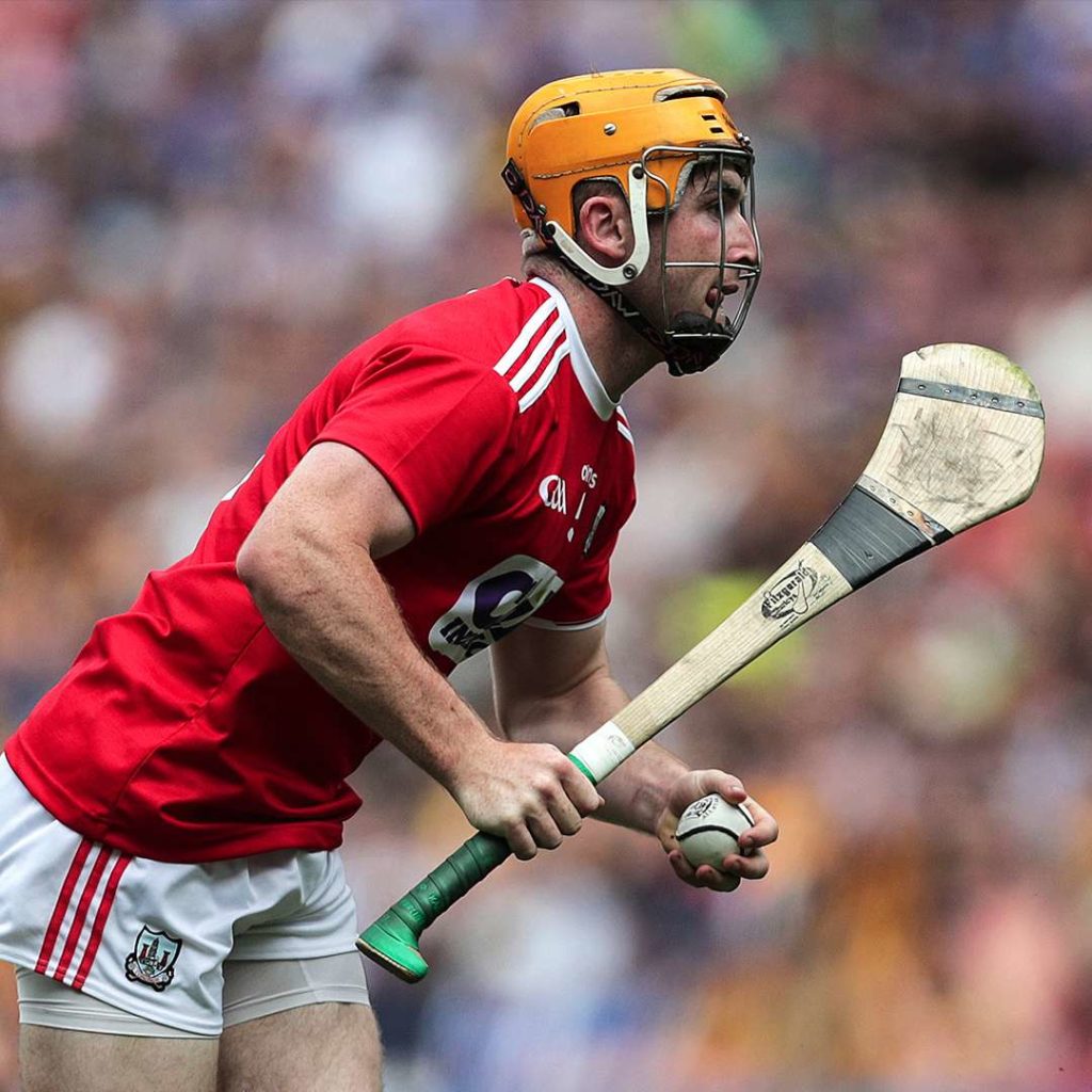 Hurling player wearing red jersey and helmet, focused with hurley and sliotar, in action during a match.