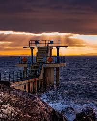Diving platform at sunset overlooking the ocean, with dramatic clouds and sunlight streaming through, creating a serene atmosphere.