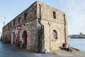 Historic stone building by the water on a sunny day with people relaxing outside, showcasing architectural charm.