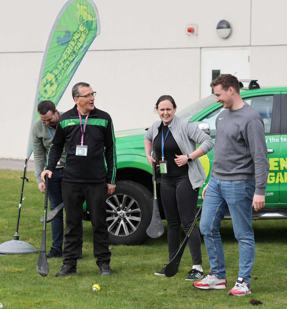 People enjoying outdoor hurling game with green banner and vehicle in the background.