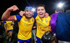 Two jubilant hurling players in yellow jerseys celebrating a victory on the field, embracing and smiling under stadium lights.