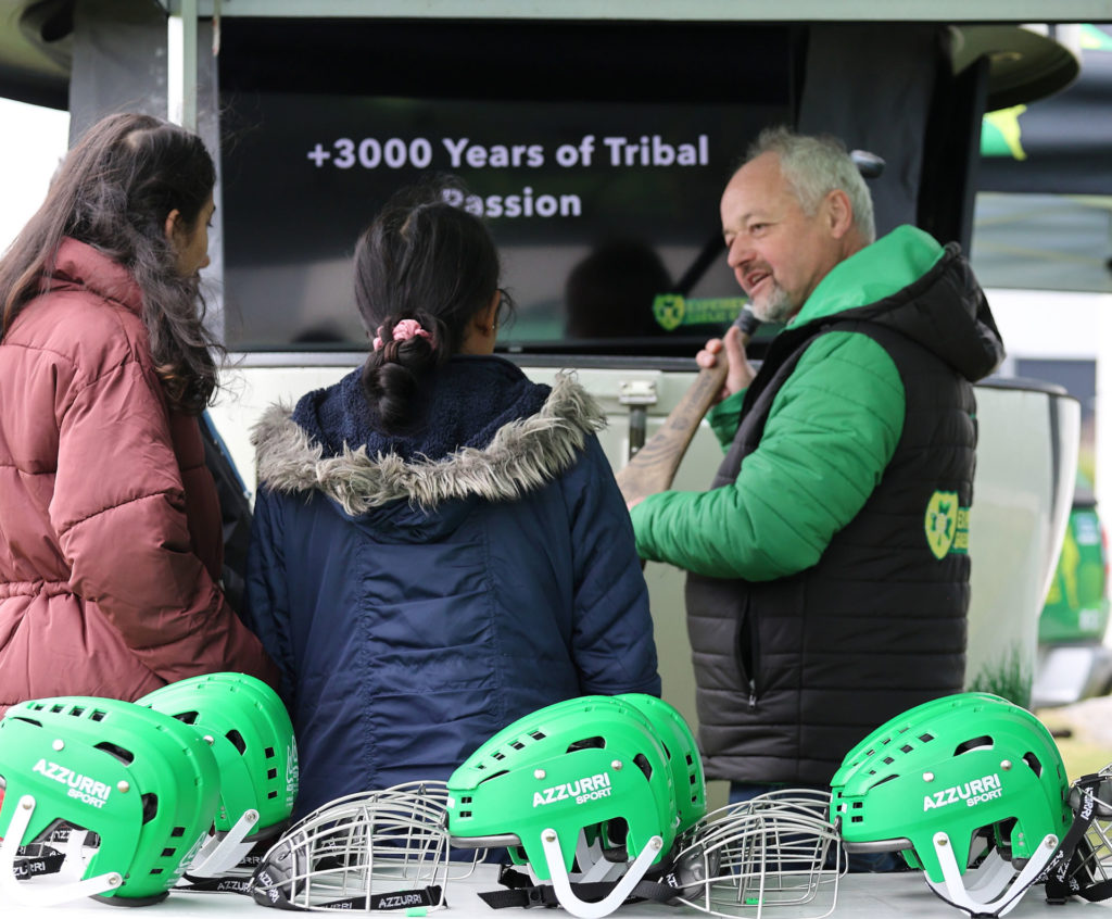 People discussing hurling equipment at an event booth, green helmets with Azzurri Sport displayed in foreground.