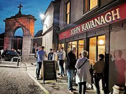 People gather outside John Kavanagh pub near a lit archway at dusk, creating a lively evening scene.