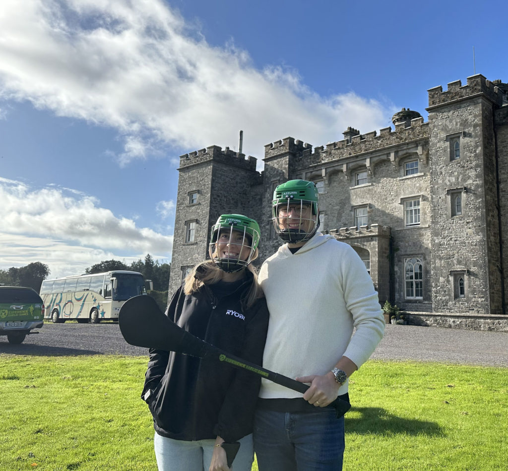 Two people in helmets with hurling sticks standing in front of a historic castle under a blue sky.