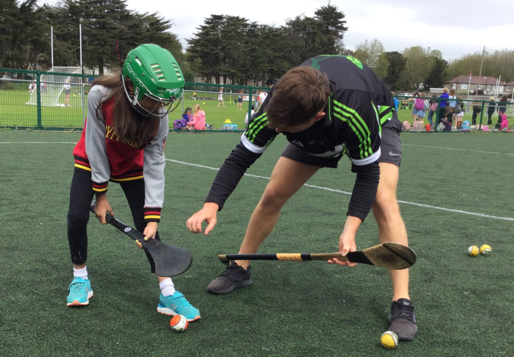 Child learning hurling with coach on a sports field, wearing helmets and jerseys, holding hurleys near sliotar.